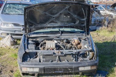 Tozlu bir motor bölmesi ve kayıp farlar için kaputu açık olan, kalabalık bir hurdalıkta çimenli bir arazide oturan bir siyah hatchback (VW Golf Mk3 'ü andırıyor).