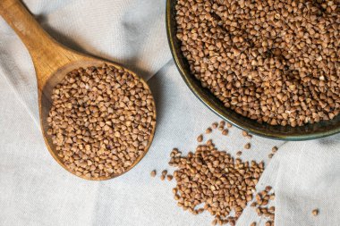 Koyu kahverengi karabuğday dolu bir seramik kâse, daha fazla tahıl içeren bir tahta kaşığın yanında yetişiyor, bej bir keten kumaş üzerine yerleştirilmiş, kırmızı böğürtlen desenli..