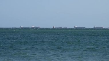 Uzak deniz ufkunda, berrak, soluk mavi gökyüzünün altında demirli büyük kargo gemilerinden oluşan bir hat..