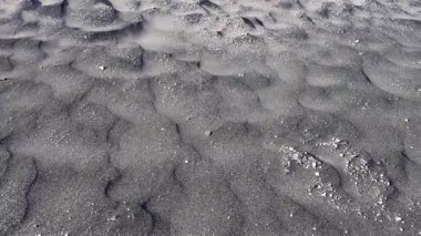 Rüzgarlı bir sahilde, küçük beyaz deniz kabukları ve çakıl taşlarıyla dağılmış siyah kumların dalgalı dokusu..