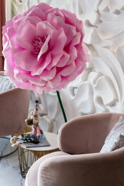 ... bej renkli kadife koltuğun yanında desenli beyaz bir duvarın karşısında duran dev bir yapay çiçek..
