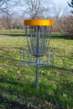 Parlak sarı bir bandı ve asılı zincirleri olan metal bir disk golf sepeti. Çimenli bir çimenlikte duruyor. Ağaçlar çıplak ve arka planda asfalt bir yol var..
