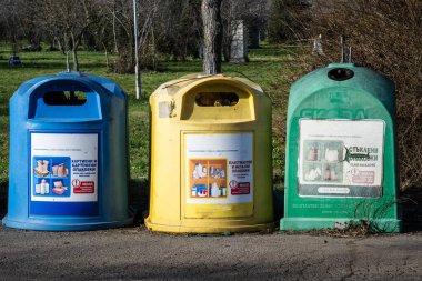 Üç sıra renkli geri dönüşüm binsblue, sarı ve yeşil bir yolda yeşil çimenlerin ve çıplak ağaçların arka planında duruyor..