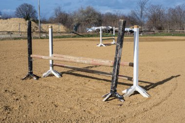 Kumlu bir arenaya kurulmuş ahşap bir gösteri atlama engeli. Dağınık bulutlarla dolu mavi bir gökyüzünün altında uzun gölgeler bırakıyor..