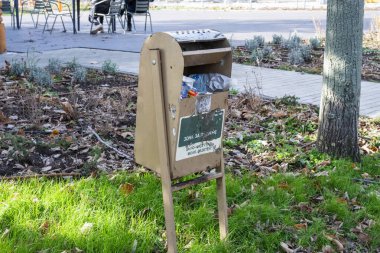 Bir ağaç gövdesi ve kaldırım yolunun yanında çimenli bir köşede duran, plastik atıkla dolup taşan, bronzlaşmış ahşap panelli bir çöp kutusu..