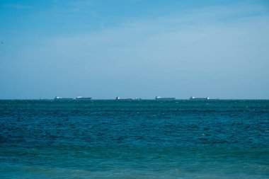 Açık gökyüzünün altında sakin mavi bir deniz. Ufukta birkaç uzak kargo gemisi var..