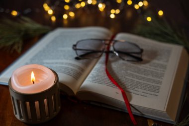 Sıcak bir mum alevi ve yumuşak arka plan peri ışıklarıyla aydınlatılmış, sayfalarda dinlenen açık bir kitap içeren samimi bir akşam sahnesi..