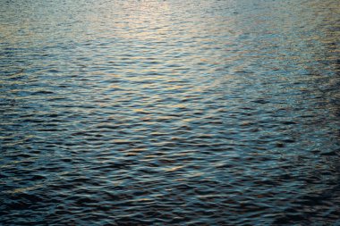 Bir su kütlesinin hafifçe dalgalanan yüzeyi, altın güneş ışığı derin mavi dalgalardan yansıyarak, parıldayan, dokulu bir desen yaratır..
