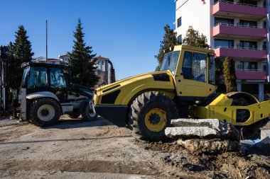 sağda büyük sarı bir yol silindiri ve solda beyaz bir traktör toprak inşaatta çalışıyor beton moloz yığınları ile bir konut binalarının zeminine karşı.