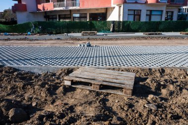 Gri geçirgen beton kaldırımlardan oluşan, ön planda gevşek toprak üzerinde duran ahşap bir palet ve arka planda bir iskan binasının yer aldığı bir inşaat alanı..