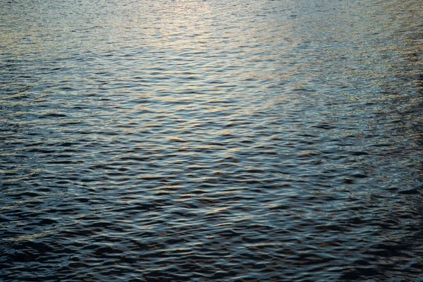 Bir su kütlesinin hafifçe dalgalanan yüzeyi, altın güneş ışığı derin mavi dalgalardan yansıyarak, parıldayan, dokulu bir desen yaratır..