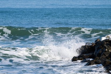 Okyanus dalgaları sağdaki koyu kayalara çarpıyor, mavi denize karşı beyaz köpük fışkırtıyor..