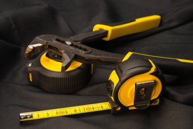 sarı ve siyah el aletlerinin bileşimi, iki geri çekilebilir bant ölçer üzerinde dinlenen ayarlanabilir bir çift pense içeren siyah kumaş arka plan.