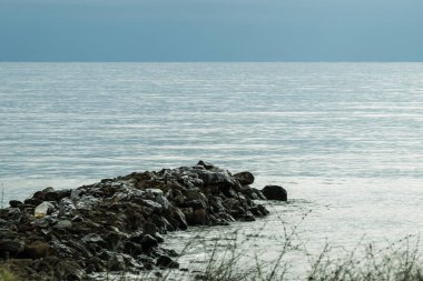 Karanlık, engebeli bir kaya iskelesi bulutlu bir gökyüzünün altında sakin, gri-mavi bir denize uzanıyor..
