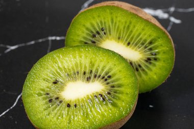 Olgun kivi meyvesinin iki yarısı koyu mermer yüzeyde canlı yeşil et, küçük siyah tohum ve beyaz çekirdek sergileniyor.