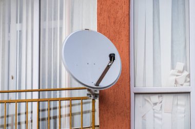 Sarı metal bir balkon korkuluğuna monte edilmiş beyaz yuvarlak uydu anteni, desenli turuncu bir duvara ve beyaz perdeli bir pencereye dayanır..