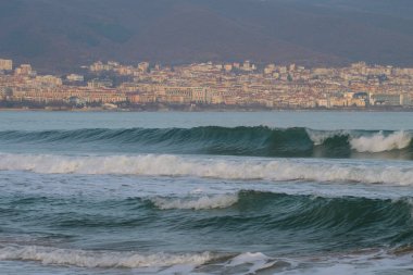 Bulgaristan 'ın Nesebar kentindeki uzak dağların eteklerine kurulmuş puslu bir kıyı şehri tarafından desteklenen, deniz mavisi yeşil dalgalar kıyıya doğru akıyor.