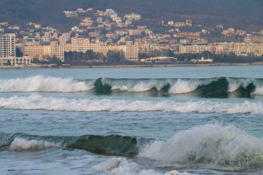 Bulgaristan 'ın Nesebar kentindeki uzak dağların eteklerine kurulmuş puslu bir kıyı şehri tarafından desteklenen, deniz mavisi yeşil dalgalar kıyıya doğru akıyor.