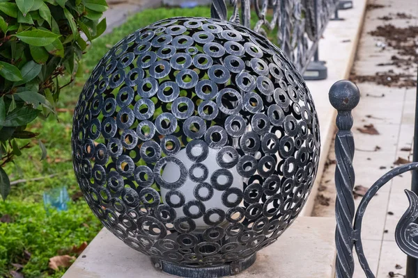Kaynak yapılmış siyah metal halkalardan yapılmış dekoratif küresel bahçe lambası, beyaz bir küre ışığına ev sahipliği yapıyor ve eğri büğrü demir bir çitin yanındaki taş duvarda dinleniyor.