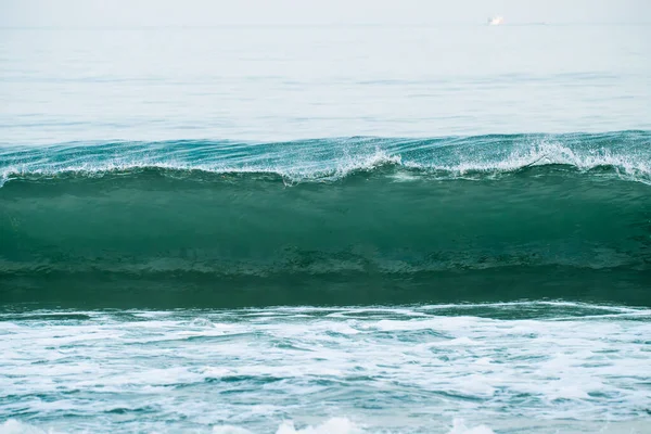 Bir deniz mavisi-yeşil dalga kıvrılıyor ve beyaz köpüklere karışıyor. Soluk, puslu bir gökyüzünün altında kıyıya yaklaşıyor..