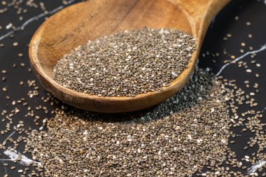 Tahta bir kaşıktan koyu dokulu bir yüzeye taşan çiğ organik chia tohumları, bu sağlıklı süper yiyeceğin benekli dokusunu vurgular..