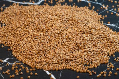 Koyu mermer bir yüzeye yayılmış çiğ karabuğday yığını, açısal şekillerini ve topraksı kahverengi tonlarını vurgular..