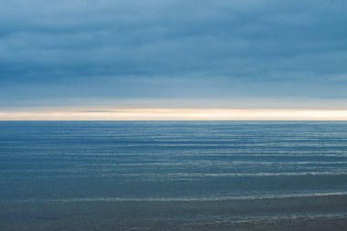 Yoğun bulutlu bir gökyüzünün altında, ufuktan yansıyan yumuşak, altın bir ışığı yansıtan ritmik yatay dalgalanmalarla derin mavi-gri okyanus engin genişliği..