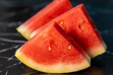 Sulu dokularını ve soluk tohumlarını vurgulamak için koyu mermer bir yüzeyde sırayla dizilmiş üç üçgen kırmızı karpuz dilimi..