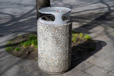 Toplu gri betondan yapılmış silindirik bir çöp tenekesi delinmiş metal kapağı ve asfalt kaldırım üzerinde şehir parkında ya da plazada bir ağacın yanında bulunan entegre kül tablası..