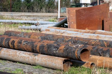 Büyük, ağır paslanmış metal borulardan oluşan bir yığın toprak ve seyrek çimenler üzerinde yatay olarak yatar ve diğer yıpranmış endüstriyel hurda malzemeler ve çelik plakalar arka planda görünür..