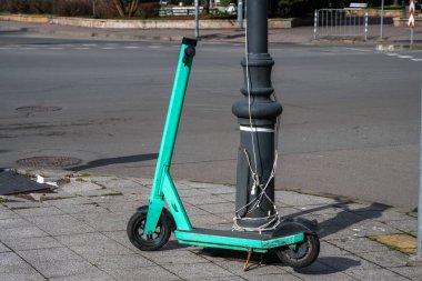 Gidonu olmayan parlak bir deniz mavisi mobilet, asfalt kavşağın yanındaki asfalt kaldırımda kablolarla birlikte koyu metalden bir sokak lambasına sıkıca bağlanmış..