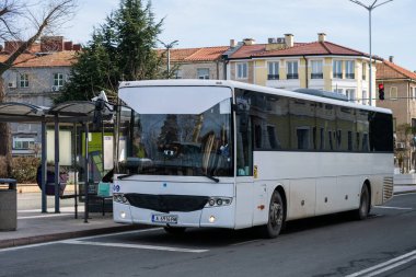 büyük beyaz bir toplu taşıma otobüsü, asfalt bir yolda, modern cam bir otobüs durağında durdu, kırmızı çatıları olan klasik Avrupa tarzı binaların arka planına karşı..