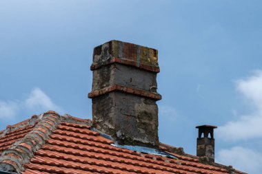 Eski, yıpranmış gri taş bir baca görünür aşınmalar ve tuğla aksanlar açık mavi gökyüzüne karşı kırmızı kiremitli bir çatıdan yükseliyor..