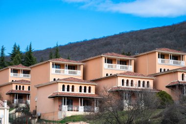 Birbirinin aynı, şeftali rengi iki katlı tatil villaları, terakotta kaplı çatıları ve beyaz balkonları var. Yoğun, ağaçlık bir tepenin arkasında parlak mavi gökyüzünün altında.