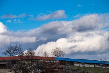 Büyük, dramatik kümülüs bulutları ve dağınık çıplak ağaçlarla dolu canlı bir mavi gökyüzüne karşı karışık kırmızı ve parlak mavi çatılı alçak kırsal tarım binasının geniş bir görüntüsü..