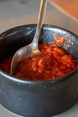 Küçük siyah bir seramik kase, dolgun, baharatlı kırmızı adjika sosu ve dinlenen metal bir kaşık..