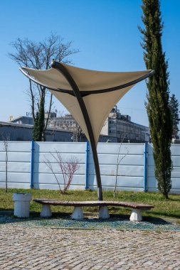 Çağdaş, ahşap bir bank. Parke taşlı bir verandada, üç noktalı mimari güneş gölgesi yapısının altında..