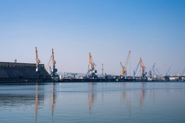 işlek bir endüstriyel liman, uzun bir sıra sarı liman vinçleri ve berrak mavi gökyüzünün altındaki sakin, cam gibi suya mükemmel yansıyan devasa beton bir yapı..