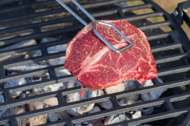 Metal maşalar, kızgın kömür ve beyaz kül üzerine kalın, baharatlı çiğ bir bifteği siyah bir ızgaranın üzerine koyar..