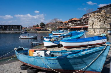 Sahilde dinlenen geleneksel mavi ve beyaz balıkçı teknelerinin bulunduğu sakin bir liman, antik taş kalıntıları ve geleneksel terasotta çatılı mimariye karşı kurulmuş.