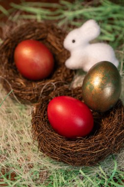 Kırmızı, koyu mor ve bronz kırmızıya boyanmış paskalya yumurtaları. Bir yumurtası örülmüş küçük bir kuş yuvasında, diğerleri ise beyaz bir paskalya tavşanı ile birlikte yeşil kağıttan bir yatakta dinleniyor.