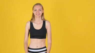 Spor kıyafetli genç sarışın kız gülümsüyor ve kopya alanı gösteriyor. Promosyon imajı için boş bir yer. Stüdyo çekimi sarı arkaplanda izole edildi.