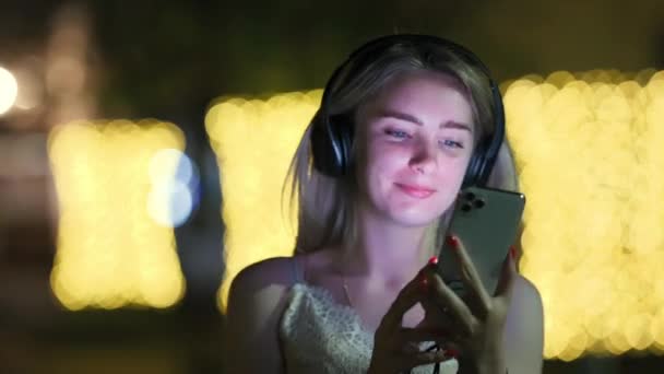 女人晚上用智能手机 在夜城的大街上 年轻姑娘的选择很可爱 她们也喜欢听音乐 — 图库视频影像