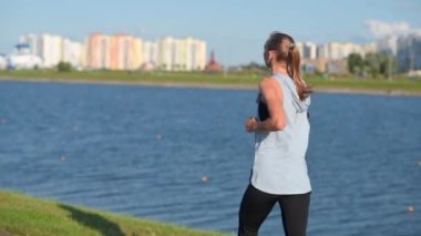 Genç kız atlet akşam parkında koşuyor. Spor Giyim 'de Formda Kız Sunset' te Nehir ve Şehrin Arkasında koşuyor. Yavaşlama Hareketi