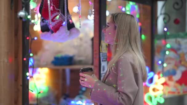 圣诞博览会上 一个穿着保暖衣服 一杯热咖啡的年轻快乐女孩的画像会选择礼物 点亮模糊的圣诞彩灯 — 图库视频影像