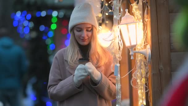 在节日城市的集市上 一个手里拿着一杯热咖啡的微笑的小女孩的画像站在圣诞灯的背景下 积极的心情快乐的情绪站在室外 — 图库视频影像