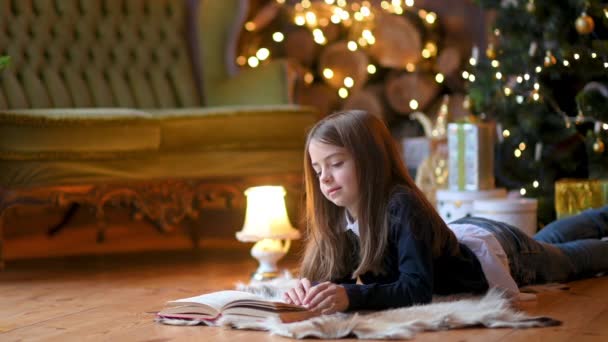 一个可爱的小女孩靠着一棵喜庆的圣诞树躺在地板上看书 背景圣诞灯饰 新年及假期概念 — 图库视频影像