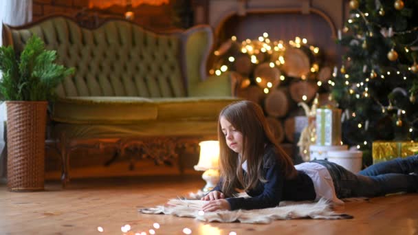 快乐的小女孩躺在地板上 靠着一棵喜庆的圣诞树 背景圣诞灯饰 新年及假期概念 — 图库视频影像