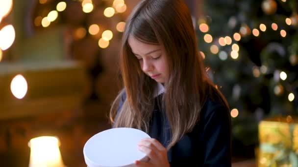 一个可爱的小女孩坐在圣诞树旁的地板上 打开礼物 快乐的女孩打开包装 打开圣诞礼物 看着盒子 假日和庆祝活动概念 — 图库视频影像
