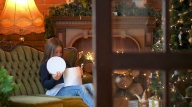 Noel ağacının yanındaki kanepede oturan tatlı küçük bir kız hediyeleri açıyor. Noel hediyesini açan mutlu kız kutuya bakıyor. Tatil ve kutlama konsepti